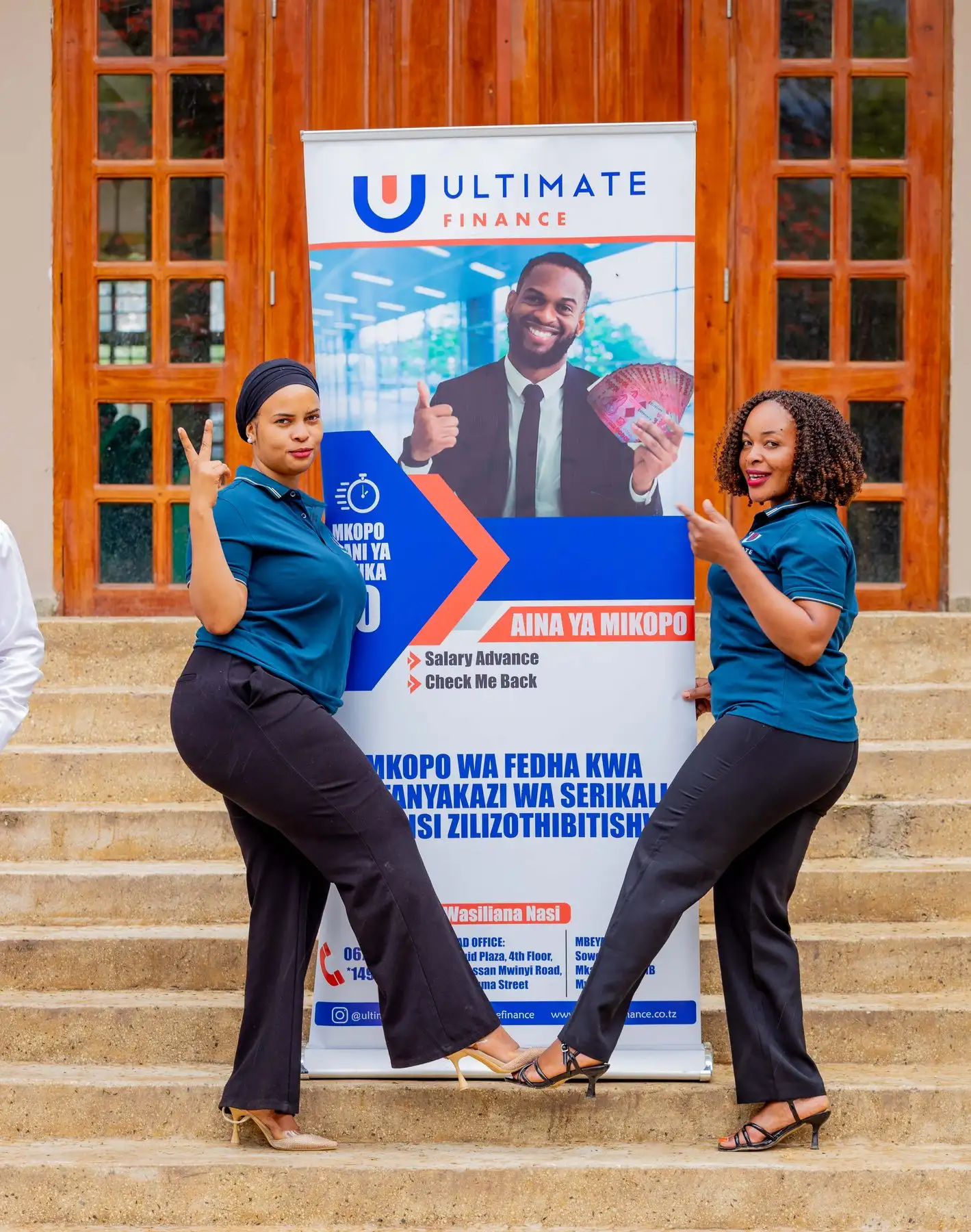 Two members of staff in branded polo shirts posing either side of an Ultimate Finance pull-up banner on office steps