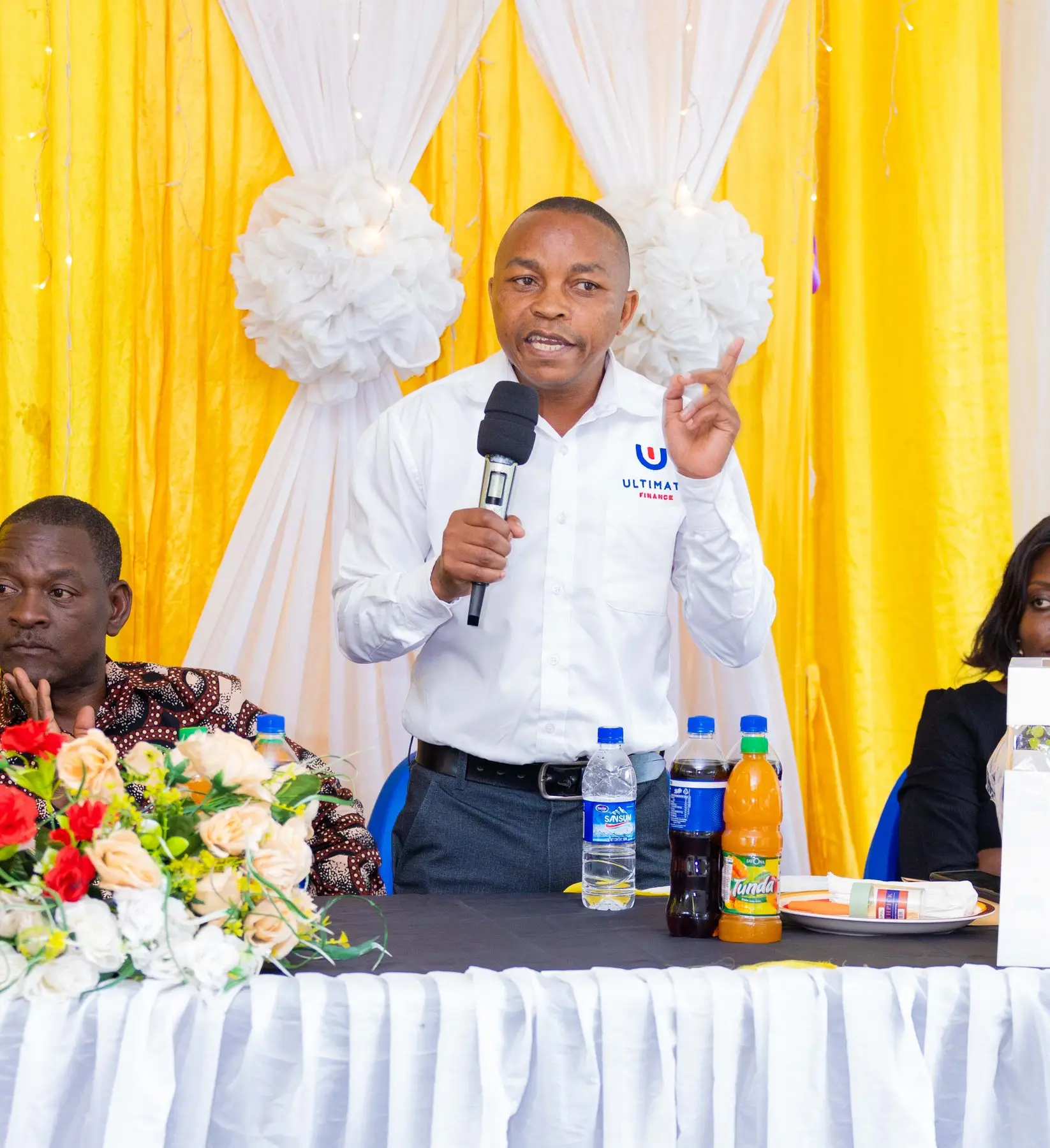 A man in an Ultimate Finance branded shirt speaking into a microphone at a customer event, seated at a table with flowers