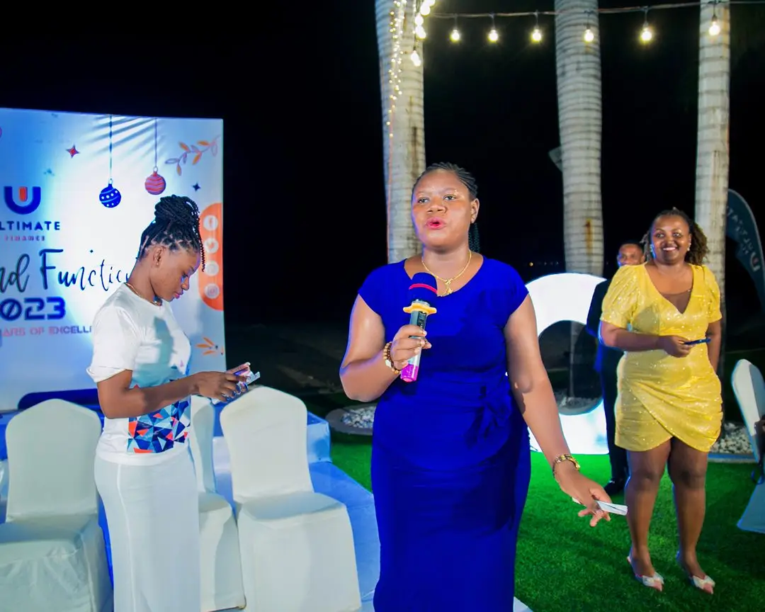 A host in a blue evening dress speaking into a microphone beside an End of Year Function 2023 backdrop