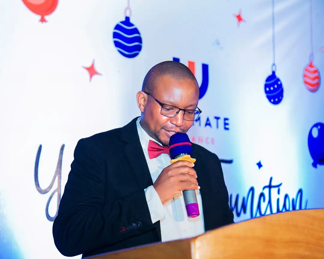 A man in a dinner jacket and bow tie speaking into a microphone at a lectern in front of a branded backdrop