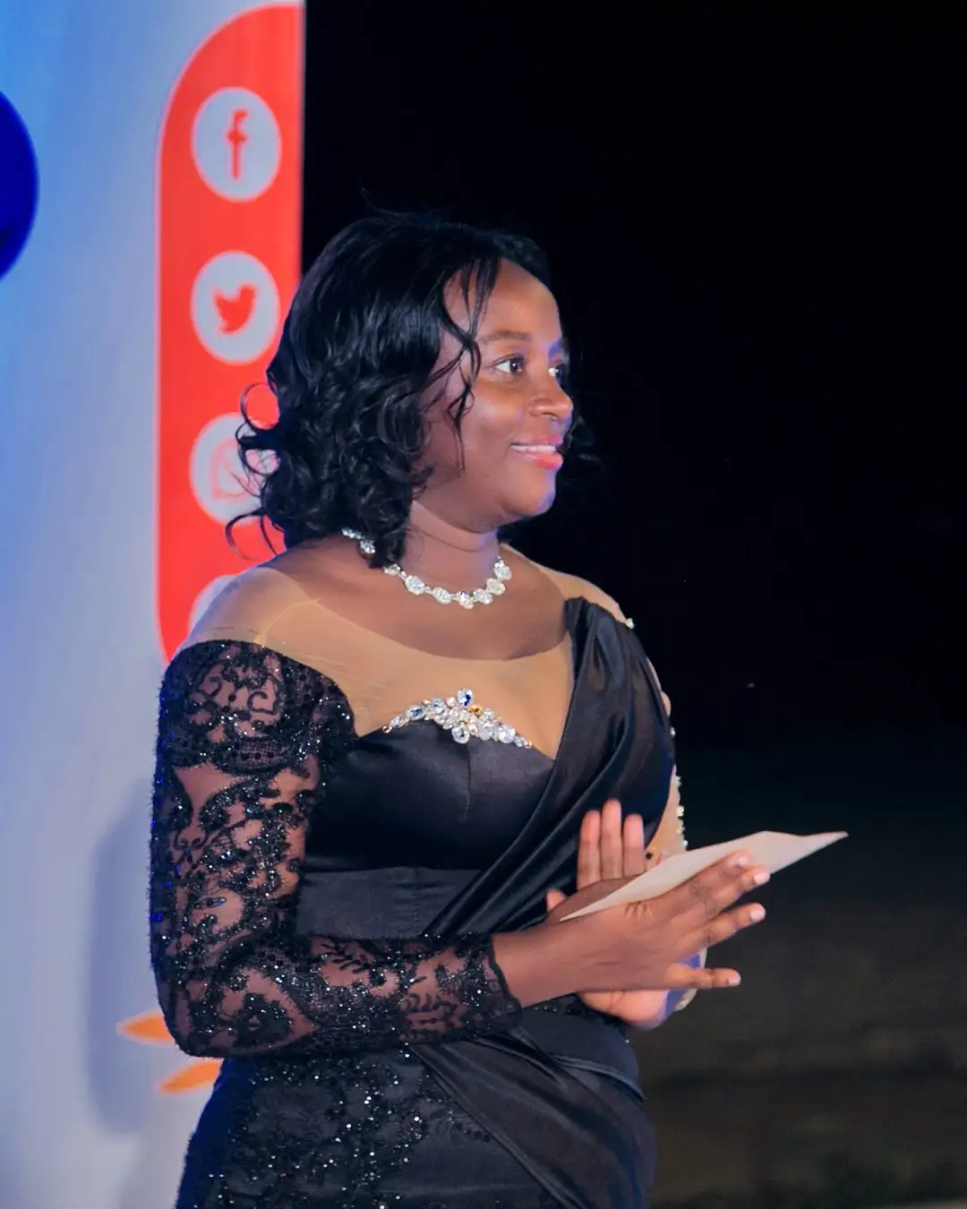 A woman in evening dress smiling and holding a certificate in front of a branded backdrop