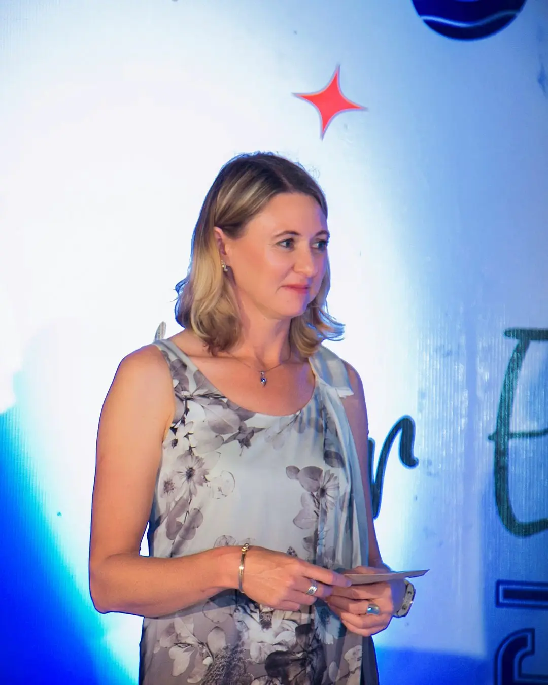 A woman in a floral dress holding a card on stage in front of the branded backdrop
