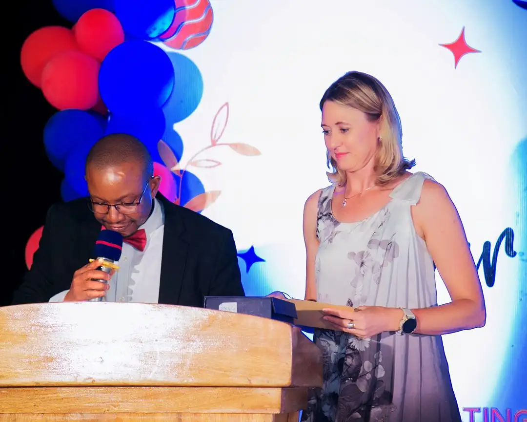 A man in a dinner jacket at the lectern with a colleague beside him holding an award, beneath a red and blue balloon arch