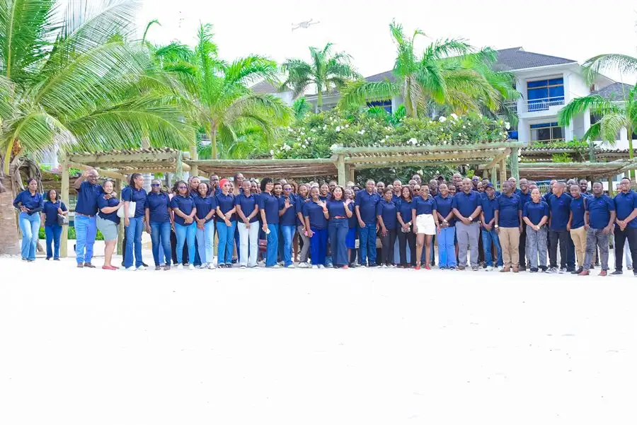 A large group photograph of the whole staff in navy branded shirts standing on sand, palm trees and buildings behind them