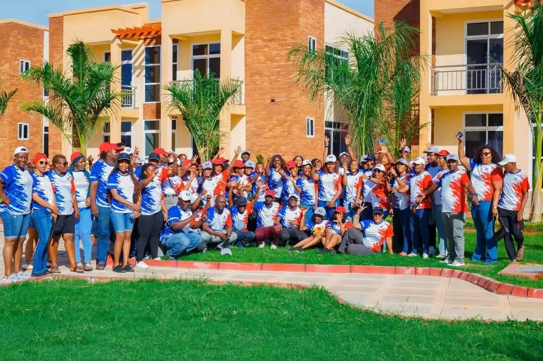A large group of staff in branded jerseys and caps assembled on a lawn in front of resort buildings and palm trees