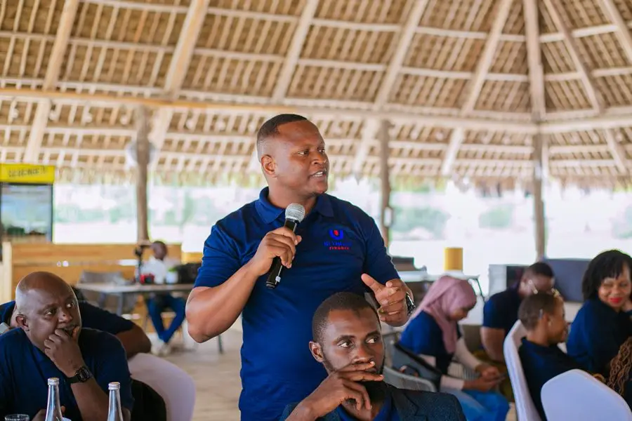 A man in a navy branded polo shirt speaking into a microphone among seated colleagues beneath a thatched roof
