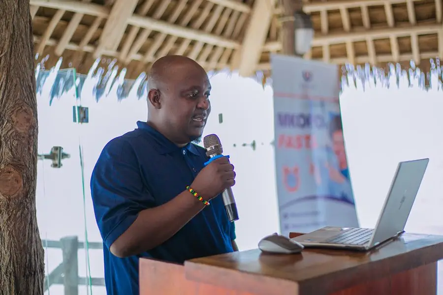 A man in a navy branded polo shirt presenting with a microphone at a lectern, beside a laptop and a branded banner