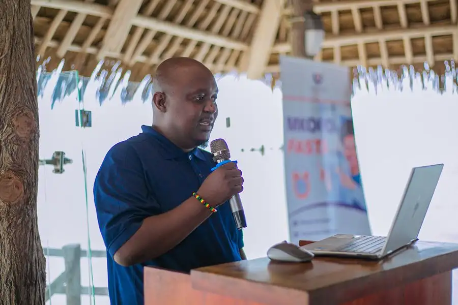 A man in a navy branded polo shirt presenting with a microphone at a lectern, beside a laptop and a branded banner