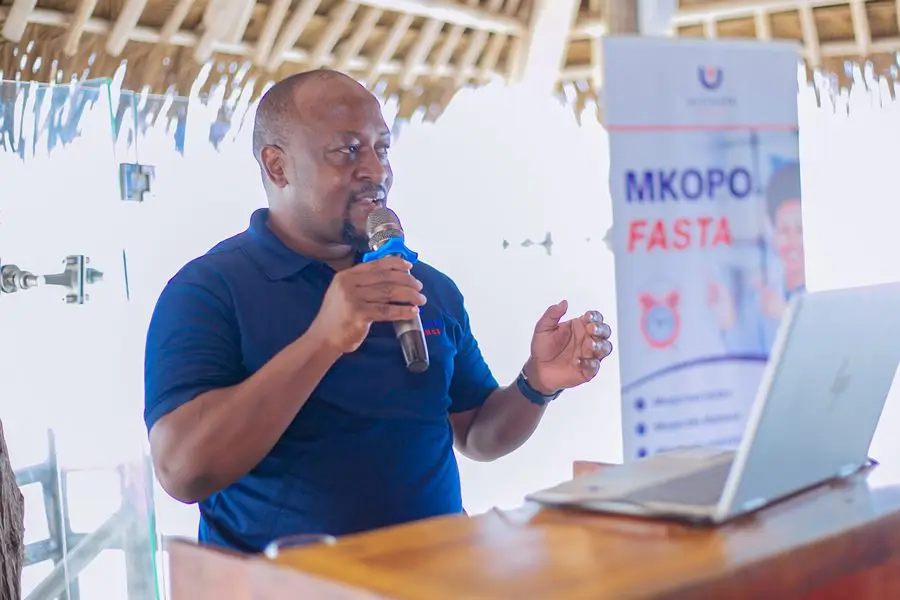 A man presenting with a microphone at a lectern beside a Mkopo Fasta product banner, beneath a thatched roof