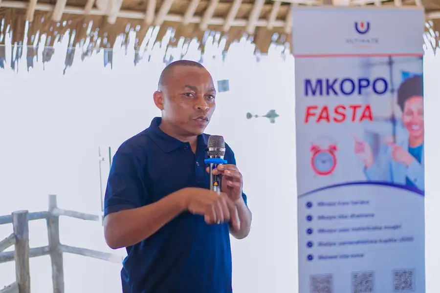A man in a navy branded polo shirt speaking into a microphone beside a Mkopo Fasta product banner