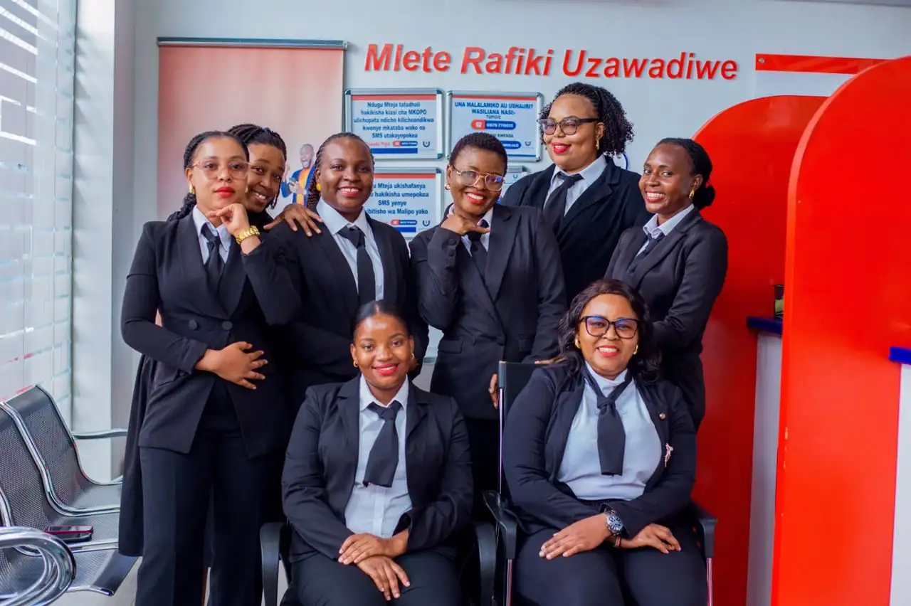 Eight members of staff in dark suits posed in a branch waiting area beside the red service counter, beneath a "Mlete Rafiki Uzawadiwe" referral poster