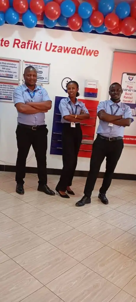 Three members of staff in pale blue branded shirts standing with folded arms beside a "Mlete Rafiki Uzawadiwe" referral poster