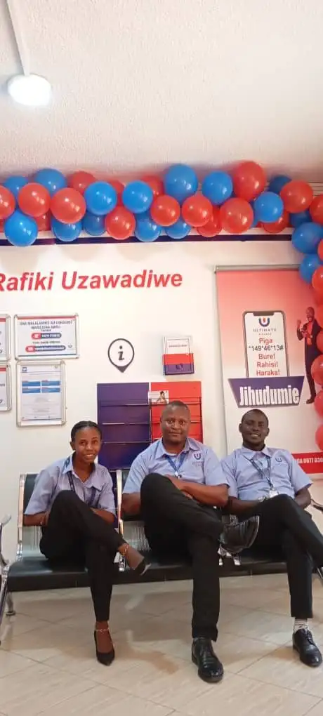 Three members of staff seated on a branch waiting-area bench beneath an arch of red and blue balloons, next to branch information posters