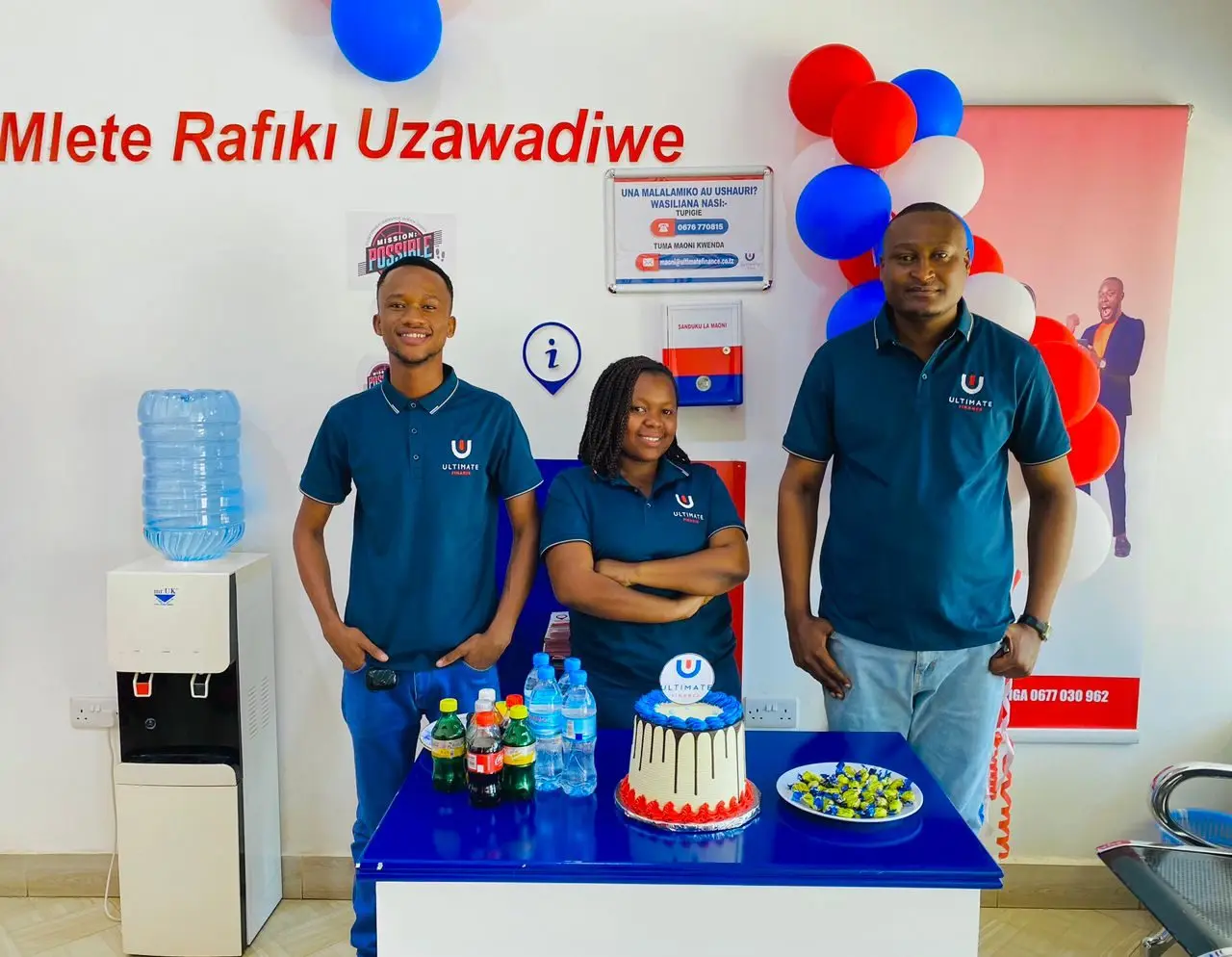 Three members of staff in branded polo shirts standing beside a table set with a decorated cake and bottled drinks, under a balloon arch