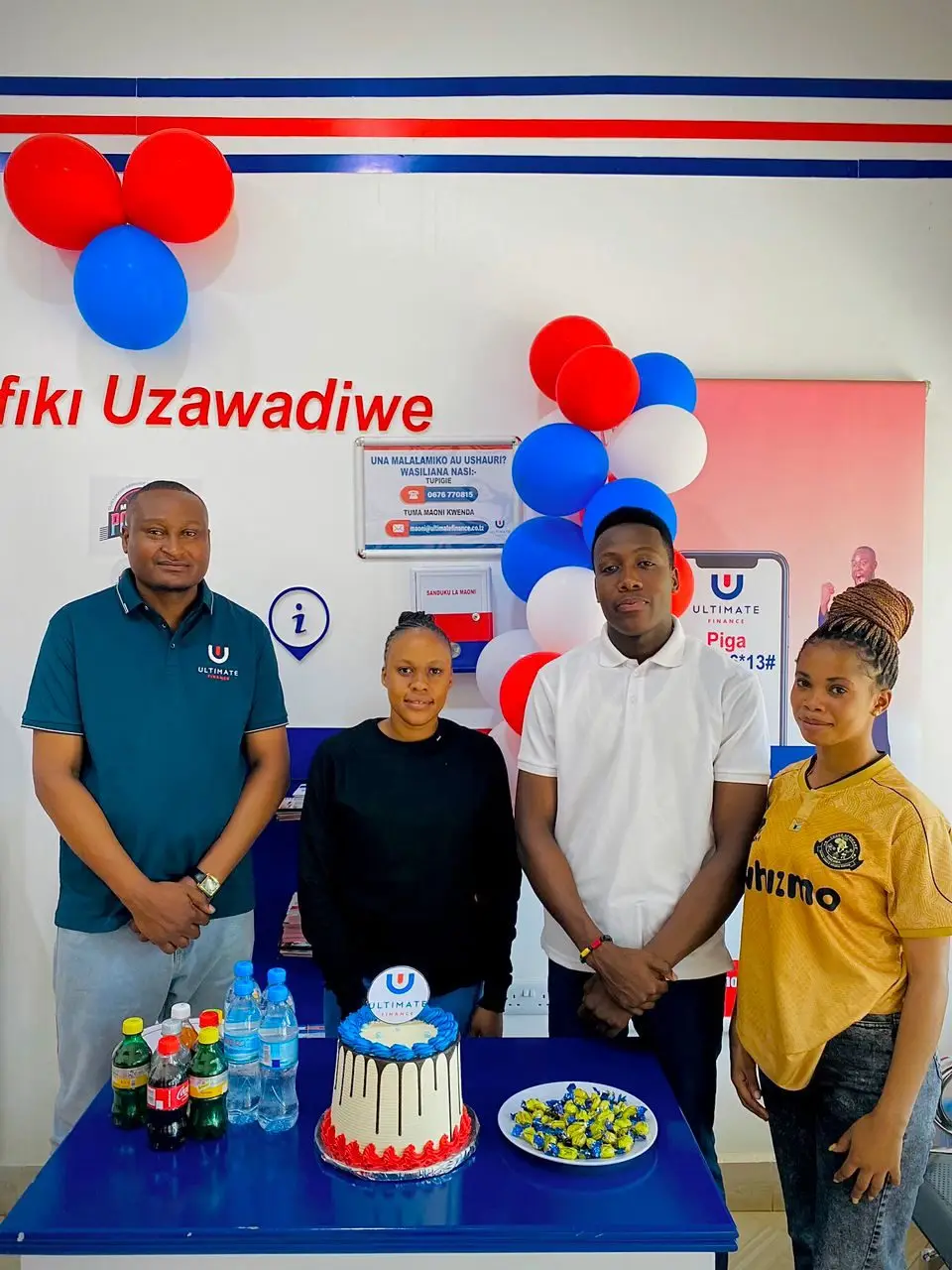 Four people standing behind a blue table holding a decorated cake and bottled drinks inside a branch
