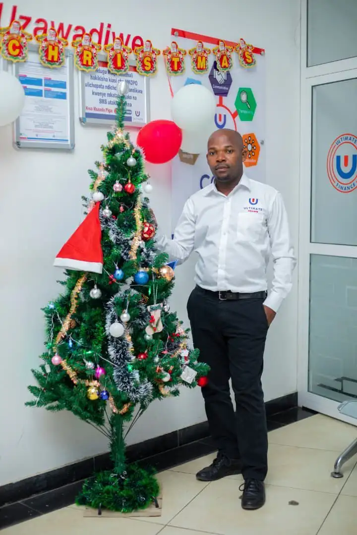 A member of staff standing beside a decorated Christmas tree with a Christmas hat hung on one branch, beneath festive bunting