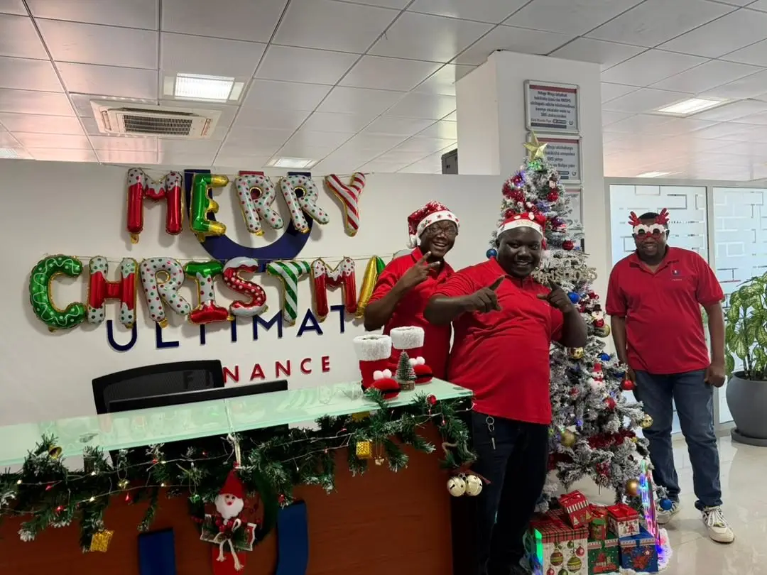 Three members of staff in front of an illuminated Merry Christmas sign mounted above the Ultimate Finance wordmark, beside a decorated tree and a garlanded counter