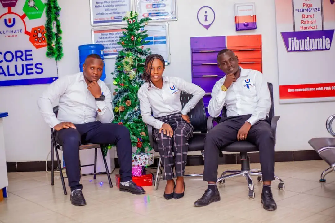 Three members of staff seated beside a Christmas tree with wrapped gifts at its base, in front of a Core Values wall panel